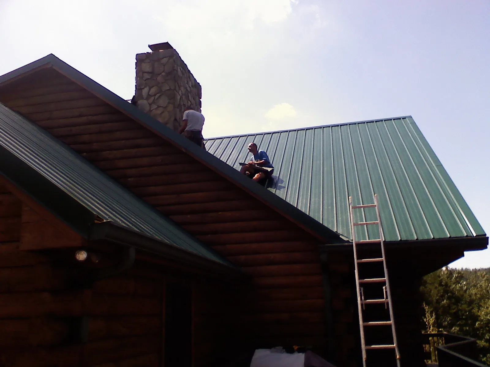 Roofers installing green metal panels beside a stone chimney on a log home in Akron